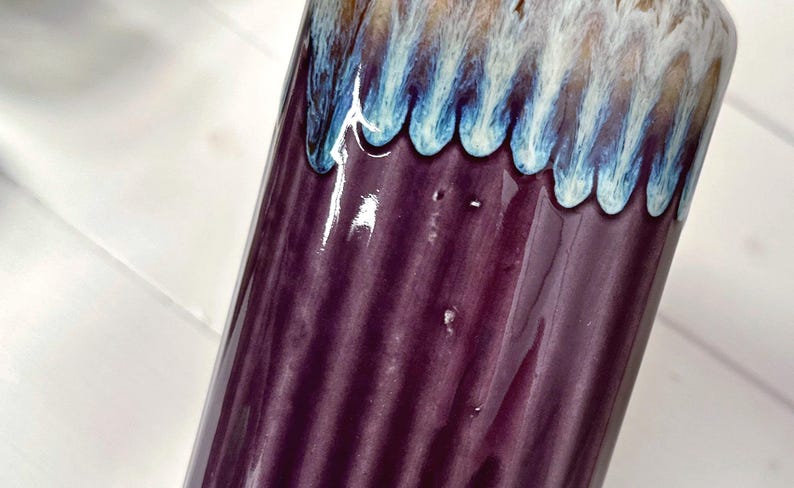 May include: A cylindrical ceramic vase with a glossy finish. The vase is primarily a deep purple color with vertical ridges. The top edge features a decorative band of blue, white, and brown glaze.