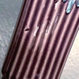 May include: Close-up of a cylindrical, dark purple object with vertical ribbed detailing. The top edge has a light blue, dripped design. The object appears to be made of ceramic or glass.