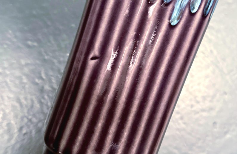 May include: Close-up of a cylindrical, dark purple object with vertical ribbed detailing. The top edge has a light blue, dripped design. The object appears to be made of ceramic or glass.