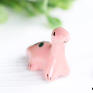May include: A small, pink ceramic dinosaur figurine with dark green spots on its back. The dinosaur has a long neck and a small head with black eyes and a black line for a mouth. The figurine is on a white surface.