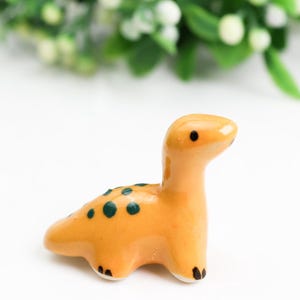 Könnte beinhalten: Eine kleine Keramik-Dinosaurierfigur in hellorange mit grünen Flecken. Der Dinosaurier steht auf einer weißen Oberfläche.