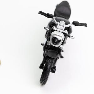 May include: A black toy motorcycle with silver handlebars and a silver headlight. The motorcycle is positioned in the center of the frame, with a coin to the left. The motorcycle's tires are black.