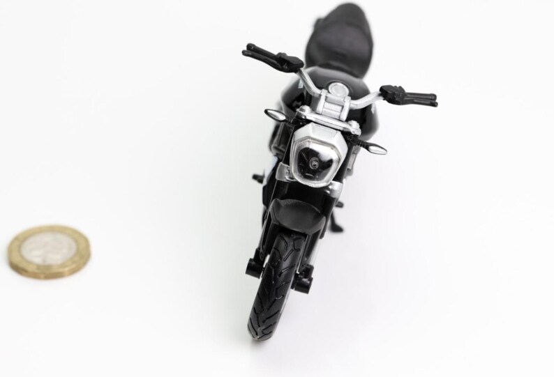 May include: A black toy motorcycle with silver handlebars and a silver headlight. The motorcycle is positioned in the center of the frame, with a coin to the left. The motorcycle's tires are black.