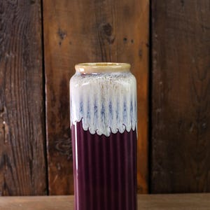 May include: A tall, cylindrical vase with a ribbed texture. The vase features a gradient of colors, starting with a deep purple at the base, transitioning to white with blue streaks, and topped with a tan rim. The vase is likely ceramic.