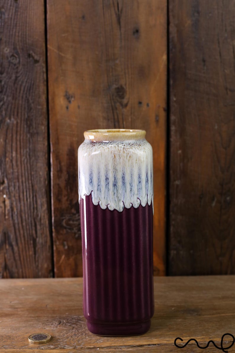 May include: A tall, cylindrical vase with a ribbed texture. The vase features a gradient of colors, starting with a deep purple at the base, transitioning to white with blue streaks, and topped with a tan rim. The vase is likely ceramic.