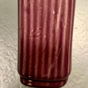 May include: A close-up of a burgundy-colored, ribbed ceramic vase. The vase has a cylindrical shape with a slightly flared base. The surface has a glossy finish. The background is a light, textured surface.