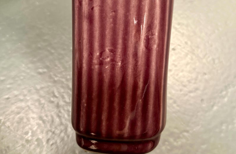 May include: A close-up of a burgundy-colored, ribbed ceramic vase. The vase has a cylindrical shape with a slightly flared base. The surface has a glossy finish. The background is a light, textured surface.