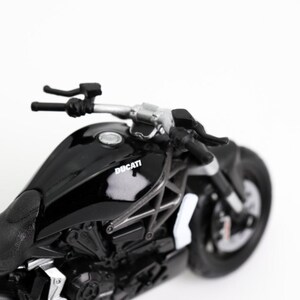 May include: A black Ducati motorcycle model with silver handlebars and a black seat. The motorcycle has the Ducati logo on the fuel tank and a black tire. The image is set against a white background.