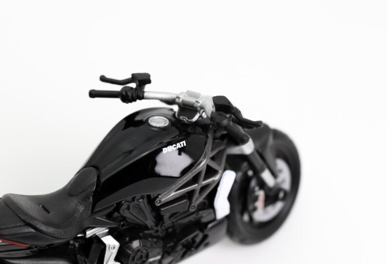 May include: A black Ducati motorcycle model with silver handlebars and a black seat. The motorcycle has the Ducati logo on the fuel tank and a black tire. The image is set against a white background.