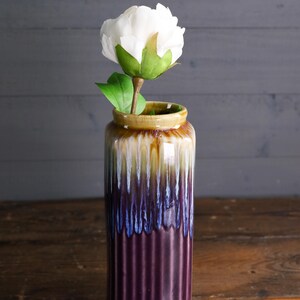 May include: A tall, ceramic vase with a white peony. The vase has a ribbed design and a color gradient of brown, yellow, blue, and purple. The flower has a green stem and leaves.
