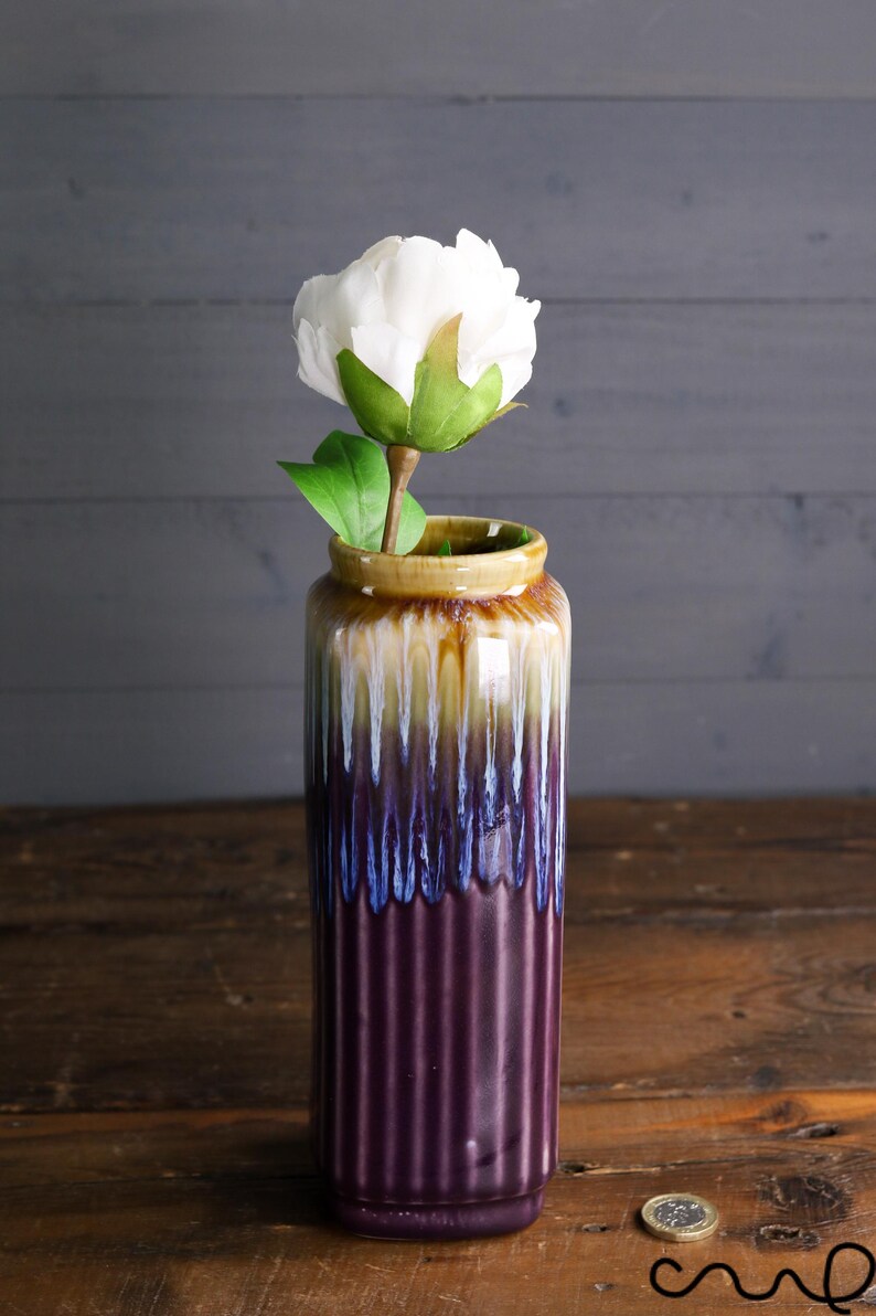 May include: A tall, ceramic vase with a white peony. The vase has a ribbed design and a color gradient of brown, yellow, blue, and purple. The flower has a green stem and leaves.