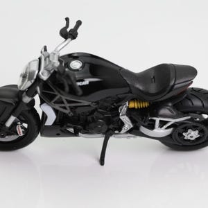 May include: A black Ducati motorcycle model with silver accents. The model features a detailed design, including the seat, wheels, and handlebars. The motorcycle is set against a white background.