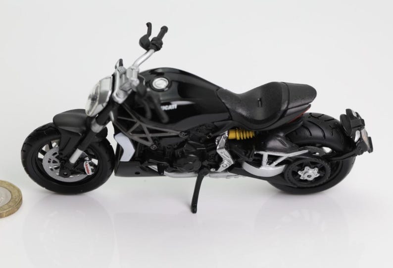 May include: A black Ducati motorcycle model with silver accents. The model features a detailed design, including the seat, wheels, and handlebars. The motorcycle is set against a white background.