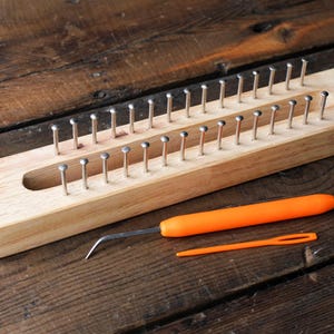 Op de afbeelding: Een rechthoekig breiraam van lichtgekleurd hout met twee parallelle rijen gelijkmatig verdeelde metalen pinnen. Inbegrepen zijn een metalen haakgereedschap met een oranje handvat en een kleine oranje plastic naald voor handwerk.