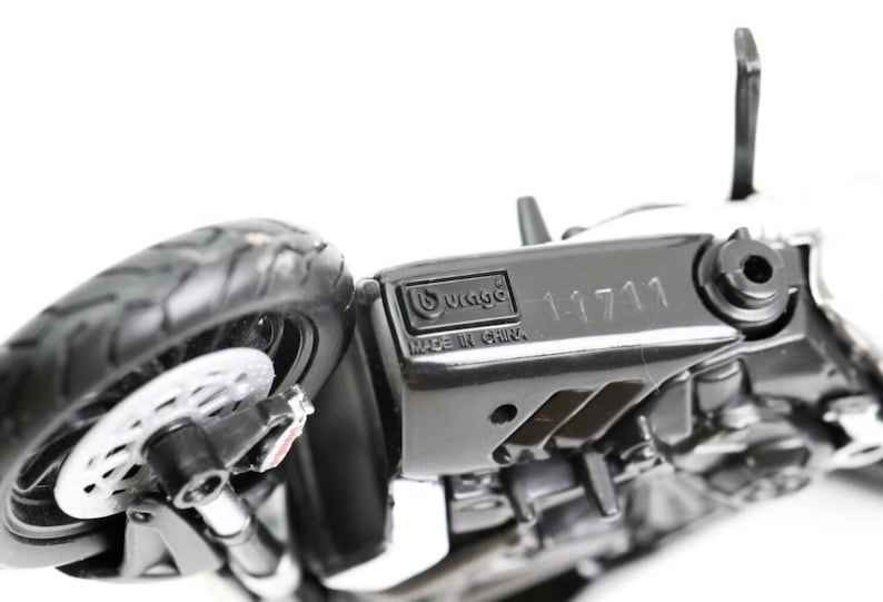 May include: A black and white toy motorcycle with a detailed design. The motorcycle has a visible tire, brake disc, and the text "11711". The brand "Burago" and "Made in China" are also visible.
