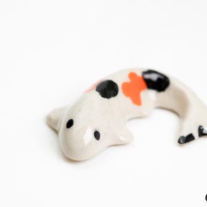 May include: White ceramic koi fish figurine with black and orange accents. The fish is curled in a swimming position.