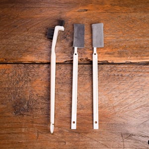 Op de afbeelding: Een set van drie reinigingshulpmiddelen met witte kunststof handvatten. Eén hulpmiddel heeft een schuine borstelkop met zwarte haren, terwijl de andere twee hulpmiddelen rechthoekige grijze stoffen pads aan de uiteinden van de stelen hebben.