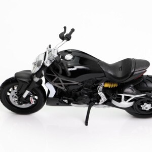 May include: A black Ducati motorcycle model with silver accents, a black seat, and yellow suspension springs. The model is on a white surface, with a coin in the foreground. The motorcycle has black tires and a kickstand.