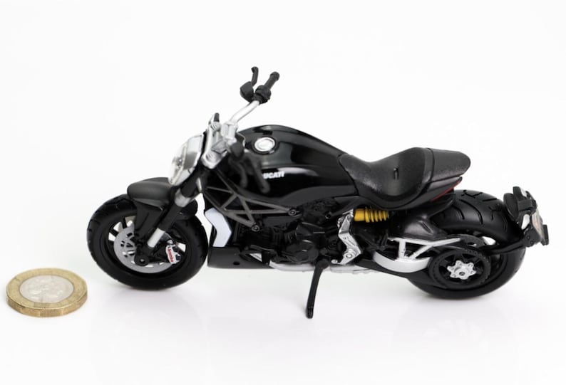May include: A black Ducati motorcycle model with silver accents, a black seat, and yellow suspension springs. The model is on a white surface, with a coin in the foreground. The motorcycle has black tires and a kickstand.