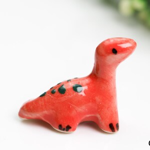May include: A small, red ceramic dinosaur figurine with black and green spots. The dinosaur is standing on a white surface.
