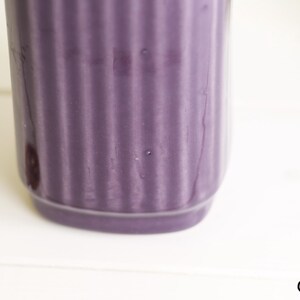 May include: A close-up of a cylindrical, purple ceramic container with vertical ribbed detailing. The container sits on a white surface, with a coin visible in the lower left corner. The container is a solid, matte purple.