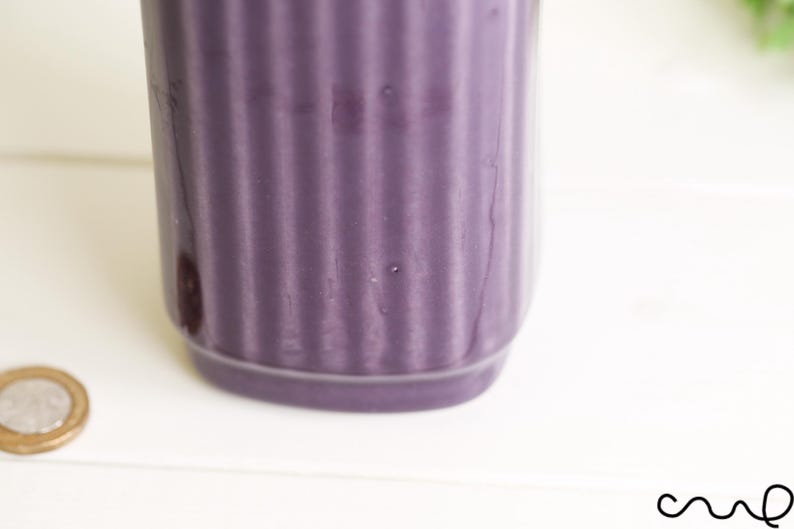 May include: A close-up of a cylindrical, purple ceramic container with vertical ribbed detailing. The container sits on a white surface, with a coin visible in the lower left corner. The container is a solid, matte purple.