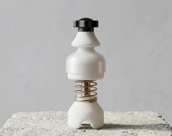 May include: A white and black salt or pepper grinder with a brass-coloured spring. The grinder has a unique design with a rounded base and a black top. It is sitting on a textured, grey surface.