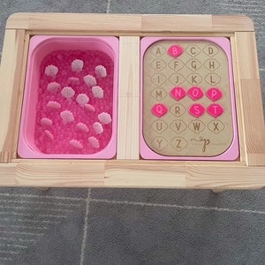 May include: A wooden activity table with two compartments. The left compartment has pink sensory beads and pink seashells. The right compartment has a wooden alphabet board with pink letters. The board says "Little Creations".