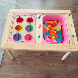 May include: A wooden activity table with a pink tray filled with colourful shapes and sticks. The table has a separate section with six circular holes labelled "Red", "Orange", "Yellow", "Green", "Blue", and "Pink".