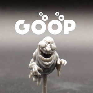 May include: A gray, stylized figurine of a smiling, one-eyed ghost-like creature, mounted on a thin, gray stand. The word "GOOP" is printed in white above the figure. The creature has a wide grin and outstretched arms.