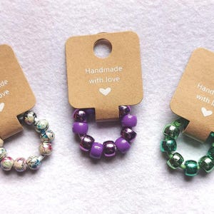 May include: Three bracelets with brown paper tags that say "Handmade with love". The bracelets are made of colourful beads. One bracelet has purple beads, one has green beads, and one has multi-coloured beads.