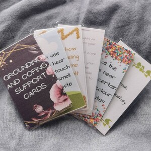 May include: A set of grounding and coping support cards with floral designs. The cards feature phrases like "see", "hear", "touch", "smell", "taste", "all the", "is near", "certain", "colour", "mindfully", "and "utes".