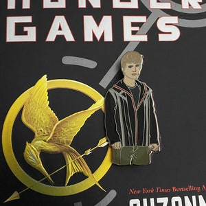 May include: A black and gold enamel pin featuring a character from the Hunger Games book series. The pin depicts a young man with short brown hair wearing a black jacket with a white stripe down the front. The pin is set against a black background with the words "The Hunger Games" printed in white.