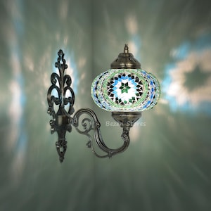 May include: A green and blue mosaic glass wall sconce with a metal frame. The sconce has a traditional design with intricate details. The light is on and illuminating the mosaic glass.