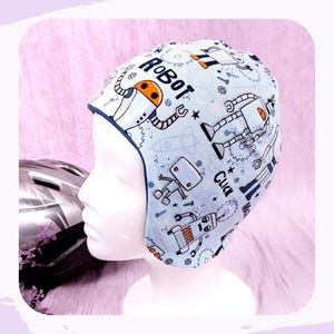 Könnte beinhalten: Hellblauer Hut mit Robotermuster und dem Wort „ROBOT“. Der Hut hat eine dunkelblaue Einfassung und Ohrenklappen. Der Hut wird auf einem weißen Schaufensterkopf präsentiert.