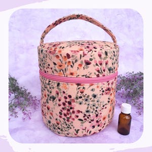 Könnte beinhalten: Eine zylindrische Tragetasche für ätherische Öle mit floralem Muster. Sie zeigt rosa, orange und grüne Blumen auf pfirsichfarbenem Hintergrund. Mit pinkfarbenem Reißverschluss und Griff. Daneben steht eine kleine braune Flasche mit weißem Deckel.