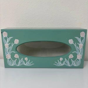 May include: Teal tissue box with an oval opening and floral accents. The box features a teal exterior with white and pink tulip designs on the corners. The tissue box is rectangular and sits on a white surface.