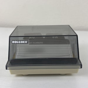 Könnte beinhalten: Ein Rolodex-Kontaktkartenordner mit einer durchscheinenden dunkelgrauen Kunststoffabdeckung und einer cremefarbenen Basis. Auf der Vorderseite befindet sich das weiße Rolodex-Logo und der Text IMPORTANT NUMBERS. Innen sind Registerkarten A-D und E-M sichtbar.