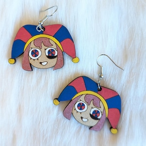 May include: A pair of wooden earrings featuring a cartoon character with pink hair, a red and blue jester hat, and black and white eyes. The earrings are on a white fuzzy background.