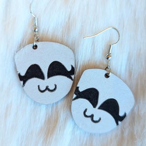May include: A pair of white wooden earrings with a black cartoon face design. The earrings have silver hooks.