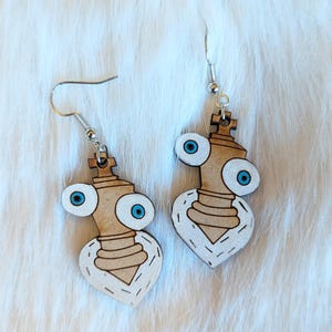 May include: A pair of wooden earrings shaped like a chess piece with googly eyes and a heart-shaped base. The earrings are painted in brown and white with blue eyes.