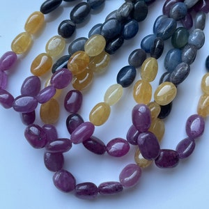 Puede incluir: Hebras de cuentas de piedras preciosas pulidas en una variedad de colores, incluyendo amarillo, morado, azul y negro. Las cuentas de forma ovalada están ensartadas, creando una exhibición texturizada y colorida. Ideal para la creación de joyas.