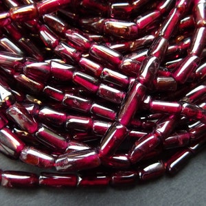 Könnte beinhalten: Eine Strähne tief roter Granatperlen, jede Perle ist ein langer, facettierter Zylinder. Die Perlen sind in einem lockeren Haufen angeordnet, wodurch die einzelnen Perlen und ihre Facetten sichtbar werden.