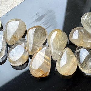 May include: A collection of clear, teardrop-shaped, golden rutilated quartz stones. The stones have a translucent appearance with visible gold inclusions.