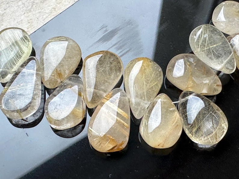 May include: A collection of clear, teardrop-shaped, golden rutilated quartz stones. The stones have a translucent appearance with visible gold inclusions.