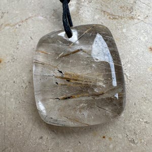 Pode incluir: Um pingente transparente, retangular com cantos arredondados, com inclusões castanho-douradas. O pingente é suspenso por um cordão preto, amarrado no topo. A pedra tem uma superfície polida e reflexiva.