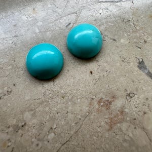 Könnte beinhalten: Zwei runde, polierte Türkis-Cabochons. Die Cabochons haben eine leuchtende, einfarbige türkise Farbe und eine glatte, gewölbte Oberfläche. Sie liegen auf einem neutralen, gesprenkelten Hintergrund.