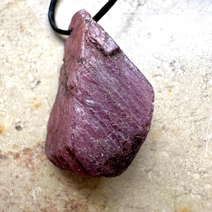Puede incluir: Un colgante de rubí en bruto. La gema de color magenta oscuro cuelga de un cordón negro. La superficie rugosa del rubí muestra líneas y texturas naturales, creando una estética única. El colgante está sobre un fondo neutro.