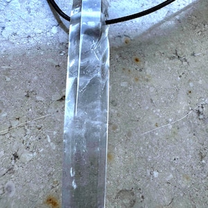 Puede incluir: Colgante de cristal de cuarzo transparente sobre un cordón negro. El cristal tiene forma rectangular con un extremo puntiagudo y una superficie texturizada. El colgante es una gema natural en bruto.