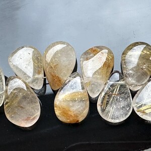 May include: A strand of teardrop-shaped beads made of clear quartz with gold inclusions. The beads are strung together with a silver wire.